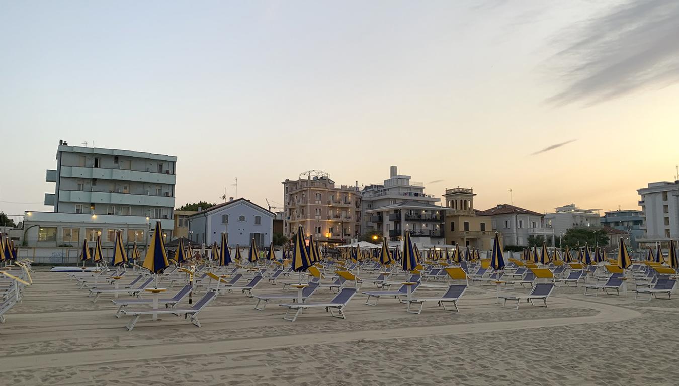 Spiaggia al tramonto con ombrelloni chiusi e edifici sullo sfondo.