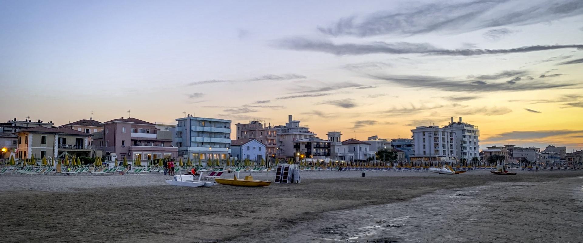 Spiaggia al tramonto con ombrelloni chiusi e edifici sullo sfondo.