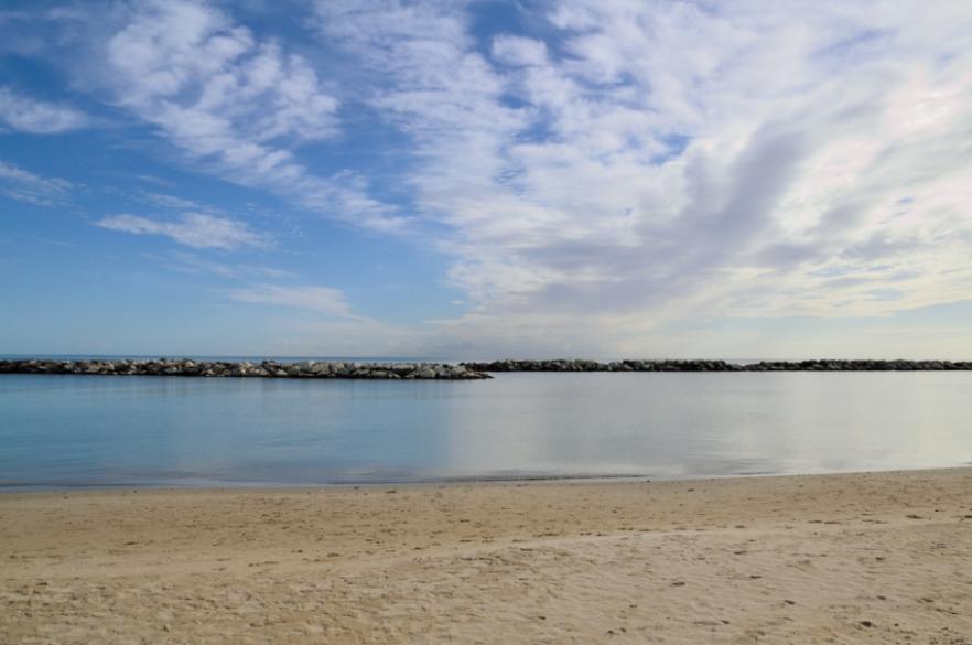 Spiaggia sabbiosa con mare calmo e cielo nuvoloso, barriera di rocce in lontananza.